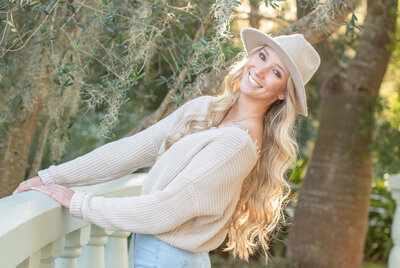 High school senior girl with long blond hair wearing a hat leaning back while holding on to a railing.