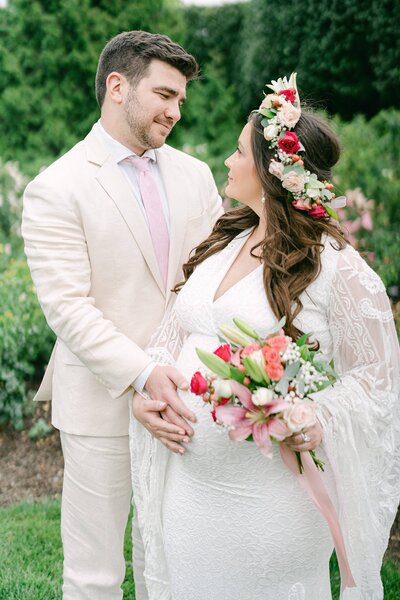 Pregnant woman in a white dress with a flower crown holding a bouquet with her husband touch her belly