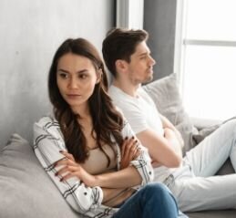 A couple with annoyed looks on their face avoid eye contact with one another. Their arms are crossed, and their body language is closed. This could symbolize a relationship that is struggling due a lack of communication. We offer communication therapy for couples in Florida. We can teach couples therapy exercises for communication