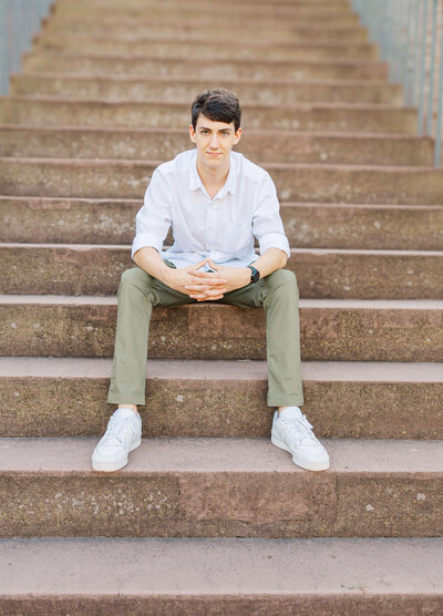 A person with short dark hair sits on wide stone steps, wearing a white shirt, olive green pants, and white sneakers. Captured by a Fayetteville NC senior photographer, they look at the camera with hands clasped in front of them, exuding a relaxed posture.
