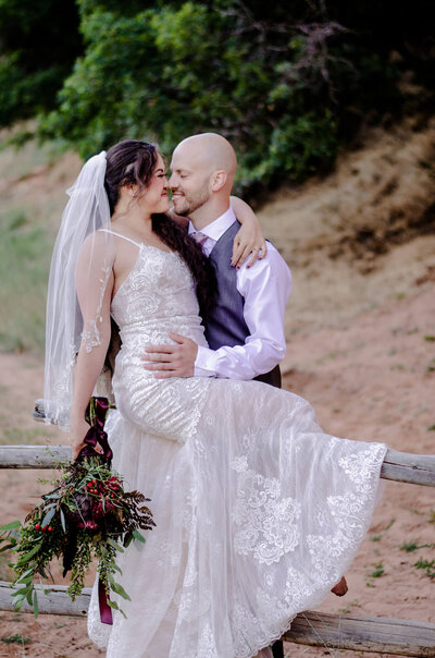 A bride holding a bouquet and sitting on a wooden fence while her groom touches noses with her