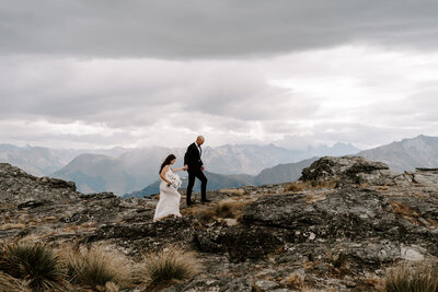 The Lovers Elopement Co - couple walk on rocks on top of mountain, heli wedding