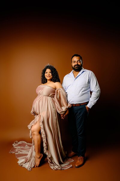 couple standing hand in hand in a maternity studio session with mom wearing a brown dress and father to be in button down shirt and pants