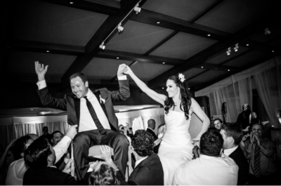 Bride and groom hold hands while being lifted in chairs at wedding reception