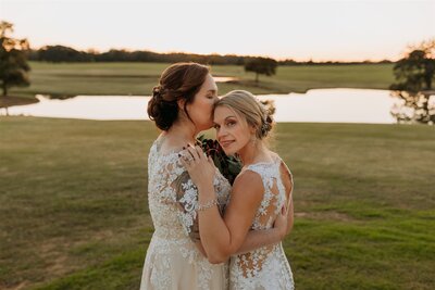 Brides hold each other closely while overlooking pond at wedding venue