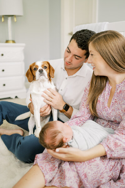 Couple smiles at King Charles Cavalier dog while holding newborn baby boy during in-home newborn photo session