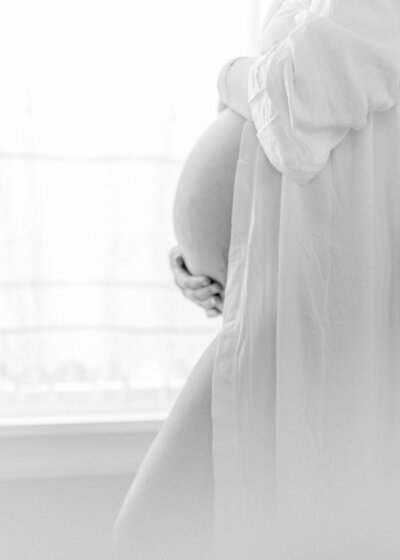 A black and white image close up of a pregnant woman holding her belly while wearing a light, white robe