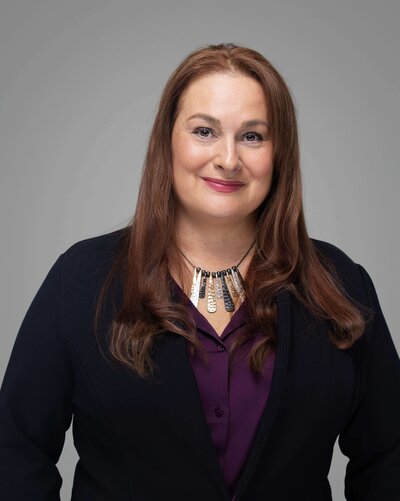 A person in a blue blazer, white t-shirt, and jeans leans against a wall outdoors. They have sunglasses on their head and their hands in their pockets. The background shows grass and a blurred landscape.