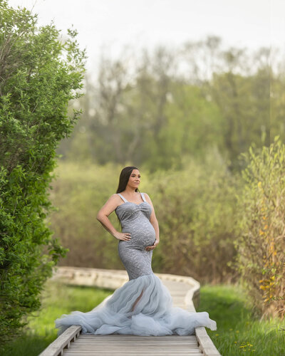A dark haired pregnant woman is wearing a long, grey, lace and tulle gown, holding her belly and standing on a wooden walkway