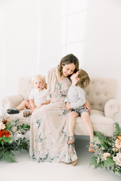 Image of mother sitting on couch with floral arrangements nearby in studio with two sons, one son kissing her cheek, taken by Sacramento Newborn Photographer Kelsey Krall