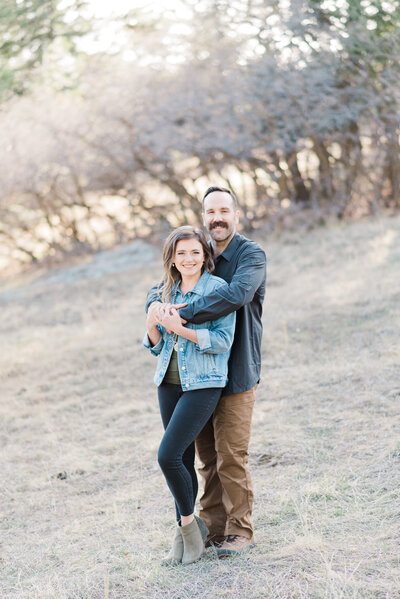 denver couples photography with man standing behind a woman and hugging her around her shoulder as she holds his hands on her chest and they both smile captured by denver engagement photographer