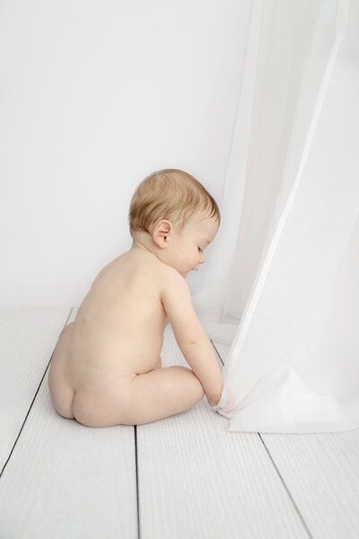little boy sits with his back to the camera during his 7 month sitter photos playing with a white curtain