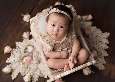 Baby girl studio milestone portrait. 6-month old Asian baby girl is wearing a cream lace romper and headband sitting in the cradle. Aerial image. Baby is looking straight at the camera with a serious expression on her face.