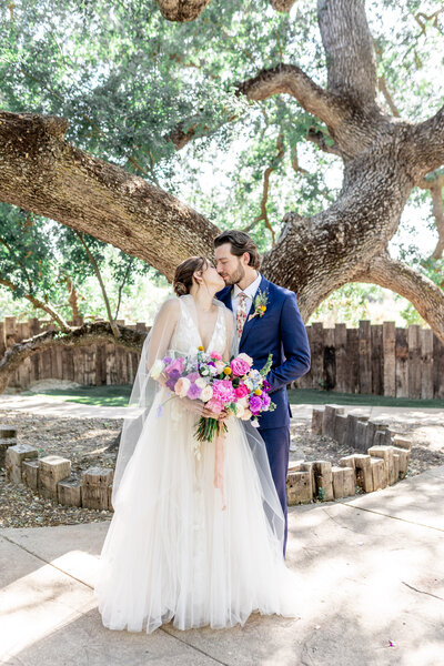 A bride and groom in springtime wedding attire kiss under the giant oak tree during their wedding at Hartley Botanica.