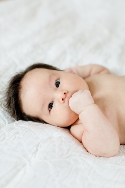 Newborn with hand in mouth looks directly at camera during newborn photoshoot.