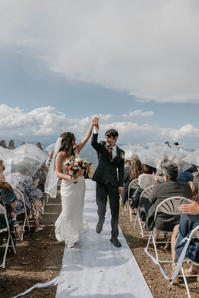 Bride and groom hold hands and celebrate walking down the aisle at an outdoor wedding with guests watching, captured in the candid style of lifestyle photography for Colorado families.