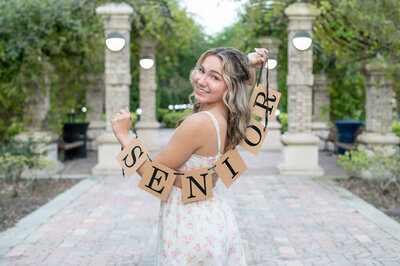 A high school senior girl holding a "Senior" sign looking back and smiling at the camera.