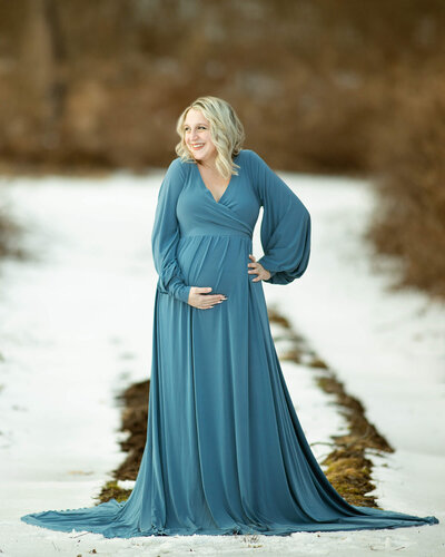 A blond woman wearing a blue maternity gown is standing in the snow, holding her belly and smiling
