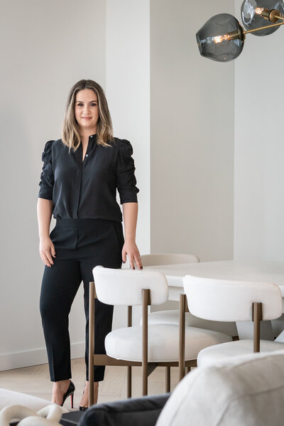 The owner Amanda Evans standing by a white dining table and white chairs, she is a white women with straight brown hair wearing black pants and a black blouse