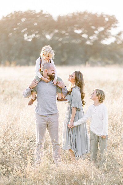 A beautiful family of 4 at a Frisco TX park standing in an open field as they are standing together and smiling.