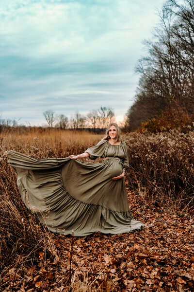 Pregnant woman wearing long flowy dress twirling her skirt in the wind surrounded by tall grasses and blue cloudy skies