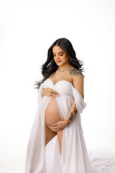 An all white themed Studio maternity image of a mother and an off-shoulder white dress with her belly exposed looking down cradling her bump