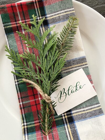 White place card with green ink calligraphy and spring of greenery on tartan plaid napkin for a holiday table