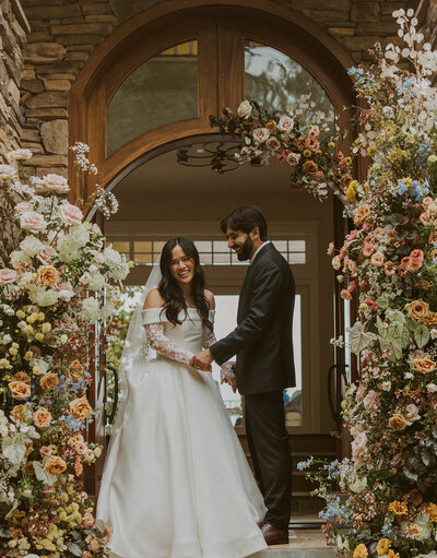Couple standing in front of floral arch outside of their home on their wedding day