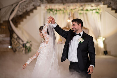 Bride and groom joyfully perform their first dance under the ornate chandelier and grand staircase at Aria Wedding