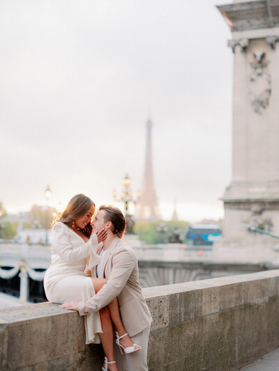 a woman in a white dress sitting on a ledge cupping a mans face who is in a tan suit with the eiffel tower and pont alexandre bridge behind them