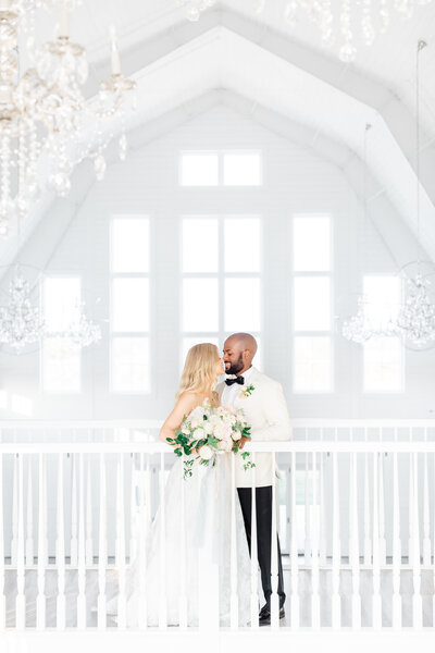 Bride and groom stand on balcony of white barn wedding venue