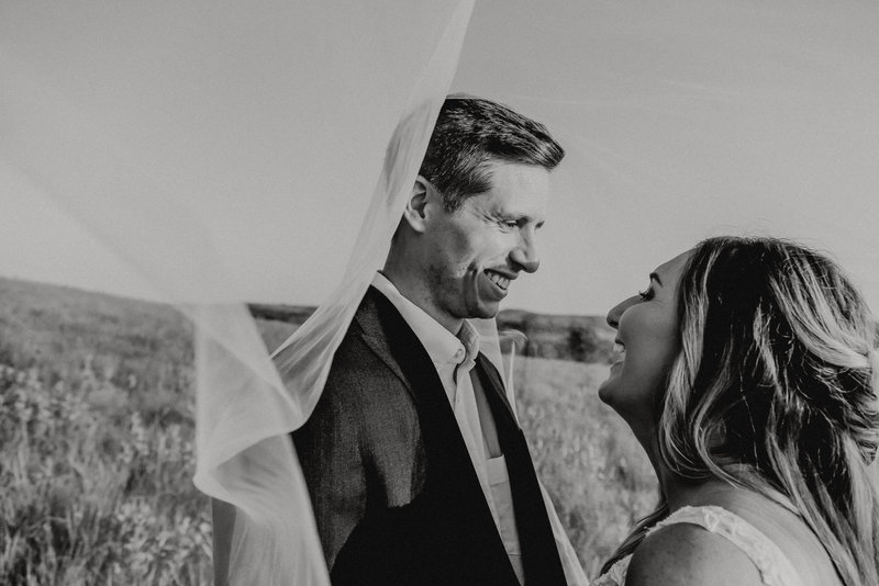 Groom and bride laughing under veil, looking at each other at summer wedding in Manhattan Kansas. Both are smiling at one another and having a good time.