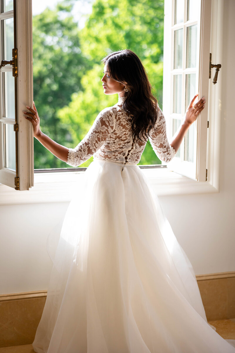 Bride dressed in a white gown with fitted lace top and long sleeves, full skirt.  Bride has opened the window shutters and the sunlight has flooded in illuminating the front of her as she looks outside.