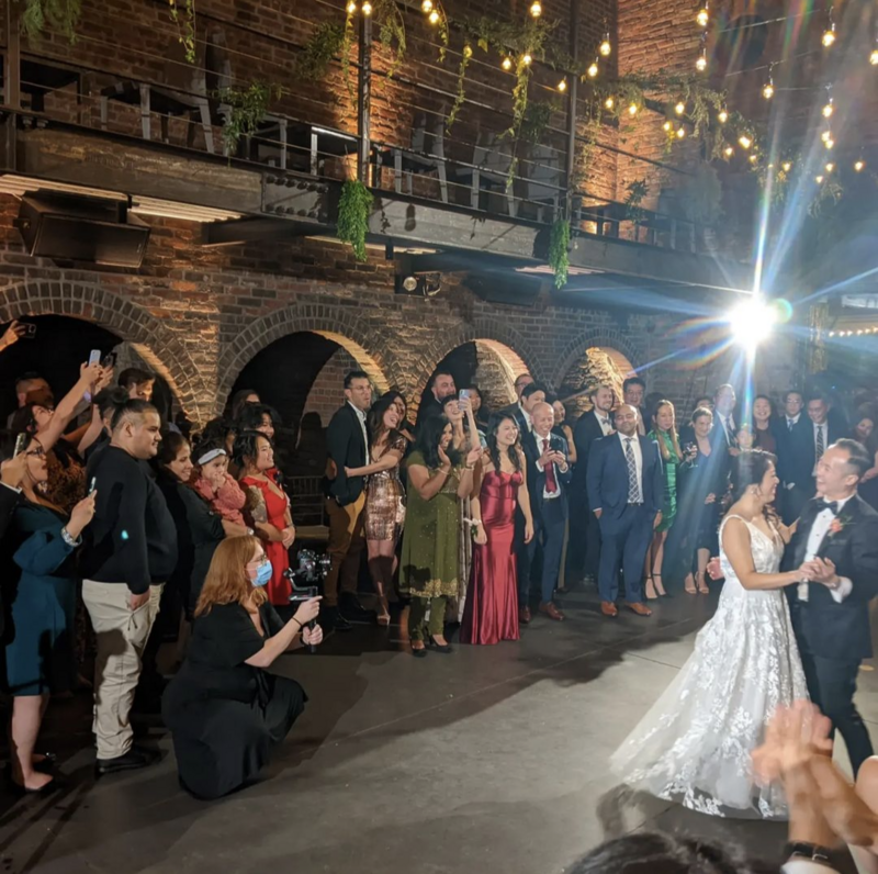A woman videographer crouching with her camera while a married couple dances and their smiling guests watch.