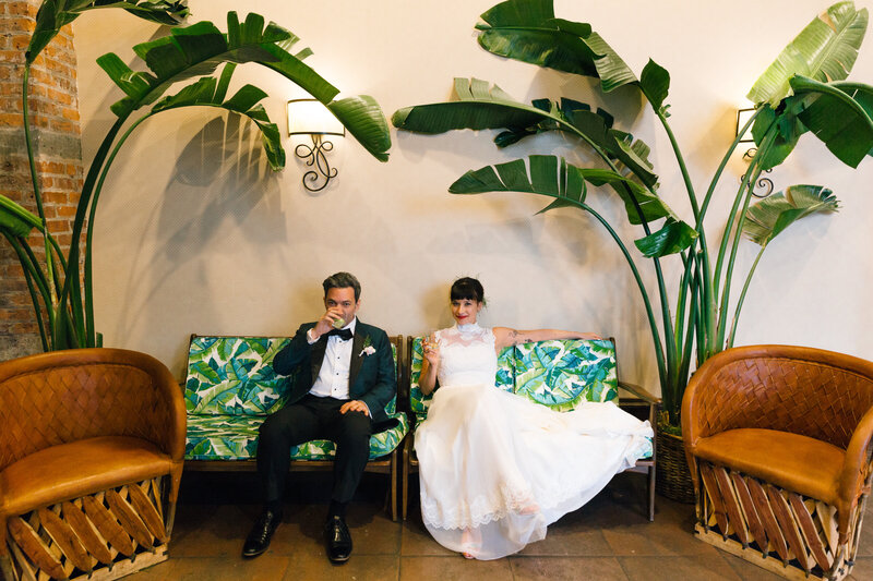 A wedding couple sitting on a couch with large plants on either side of them.