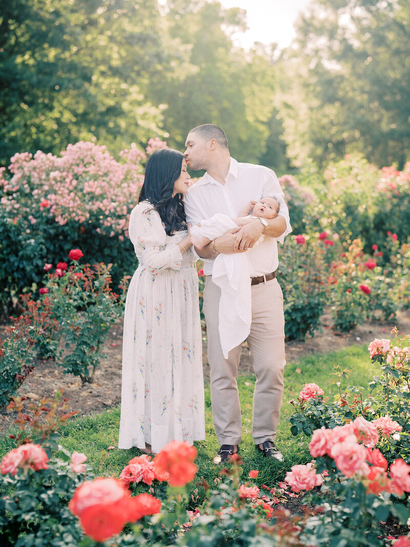 Father kisses his wife as he holds his newborn daughter in a rose garden photographed by Maryland Newborn Photographer Marie Elizabeth Photography