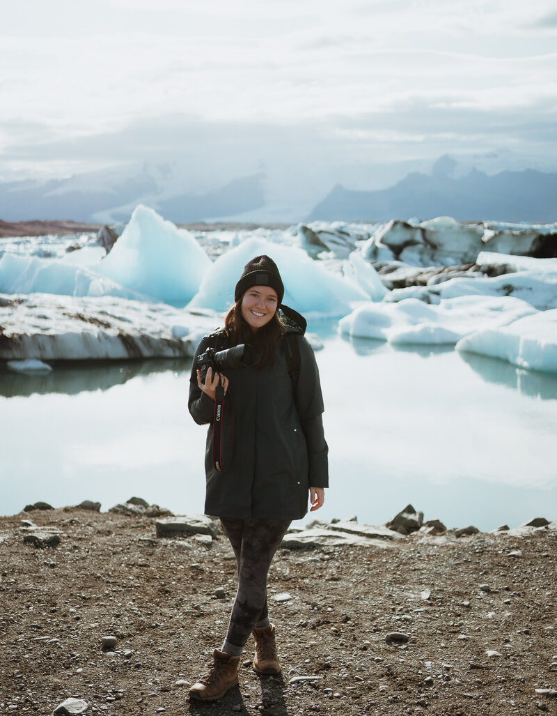 woman smiling at the camera. She is holding a camera and wearing a rain jacket along with a black beanie. She is standing in front of icebergs, and lake.