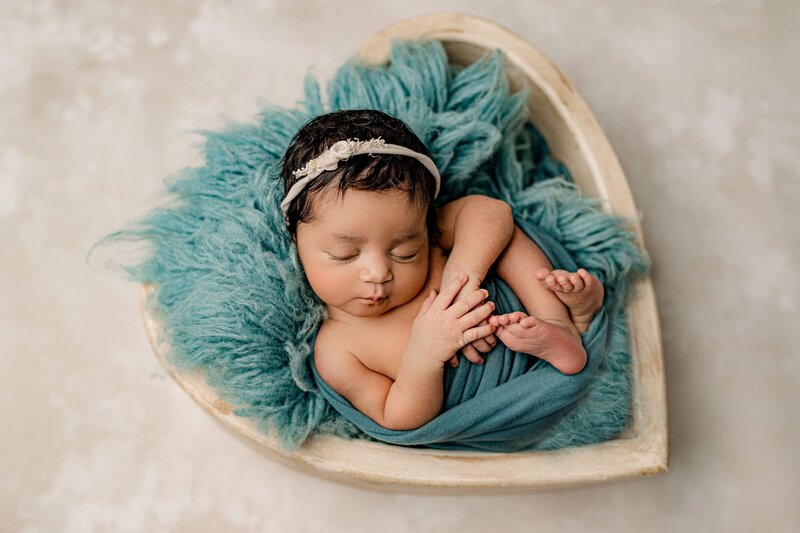 newborn baby girl is wrapped in teal and placed on a teal fur layer and a matching heart bowl with a cream bow