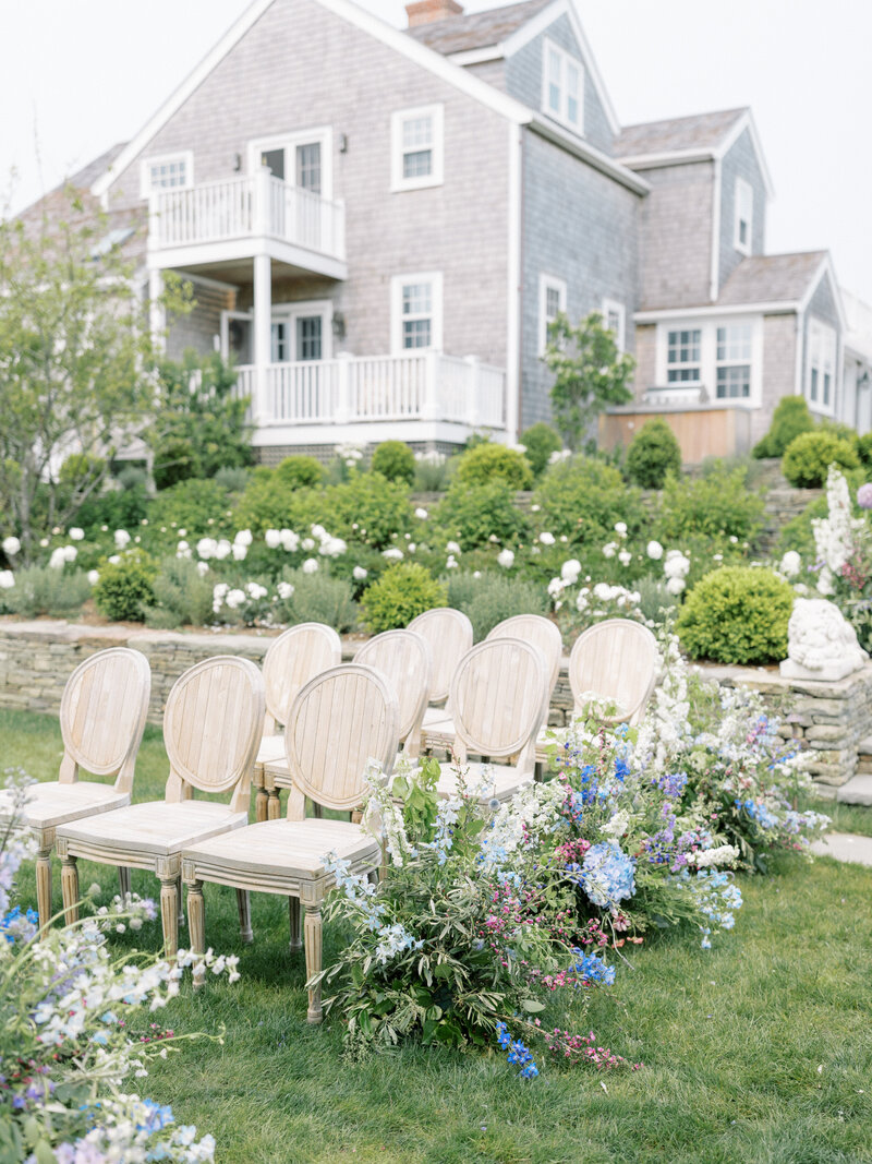 Outdoor wedding ceremony space with houses in the background and large floral arrangements on the ground along the aisle