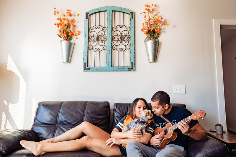 During an at-home engagement photo session a couple sits on their couch with their dog while the man plays a guitar