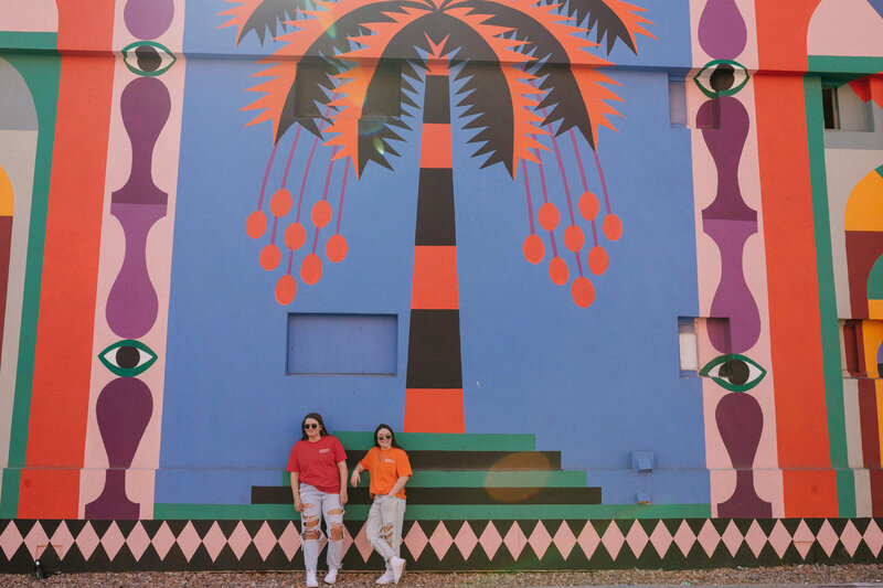 Two people leaning up against a wall with a large mural on it.