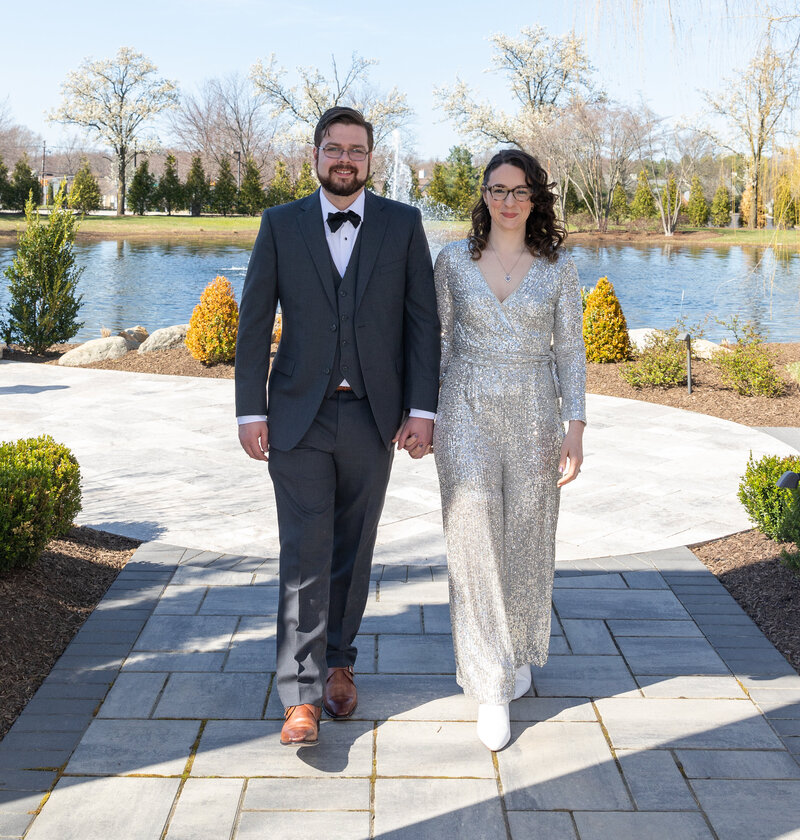 A groom and bride posing together outside by the lake. The bride has a glitter jump suit on and the groom is wearing black.