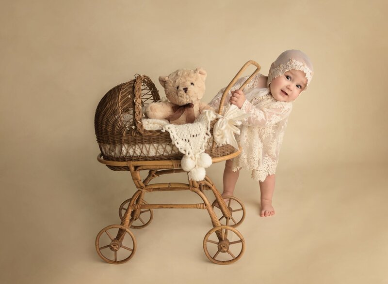 A toddler girl in a lace dress and bonnet plays with a small wicker baby carriage and teddy bear in a Lafayette Baby Milestone Photographer studio