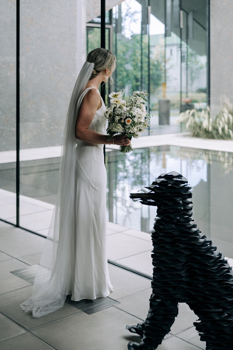 beside a sculpture of a dog a bride looks through a glass window at a pond in the christhurch cbd