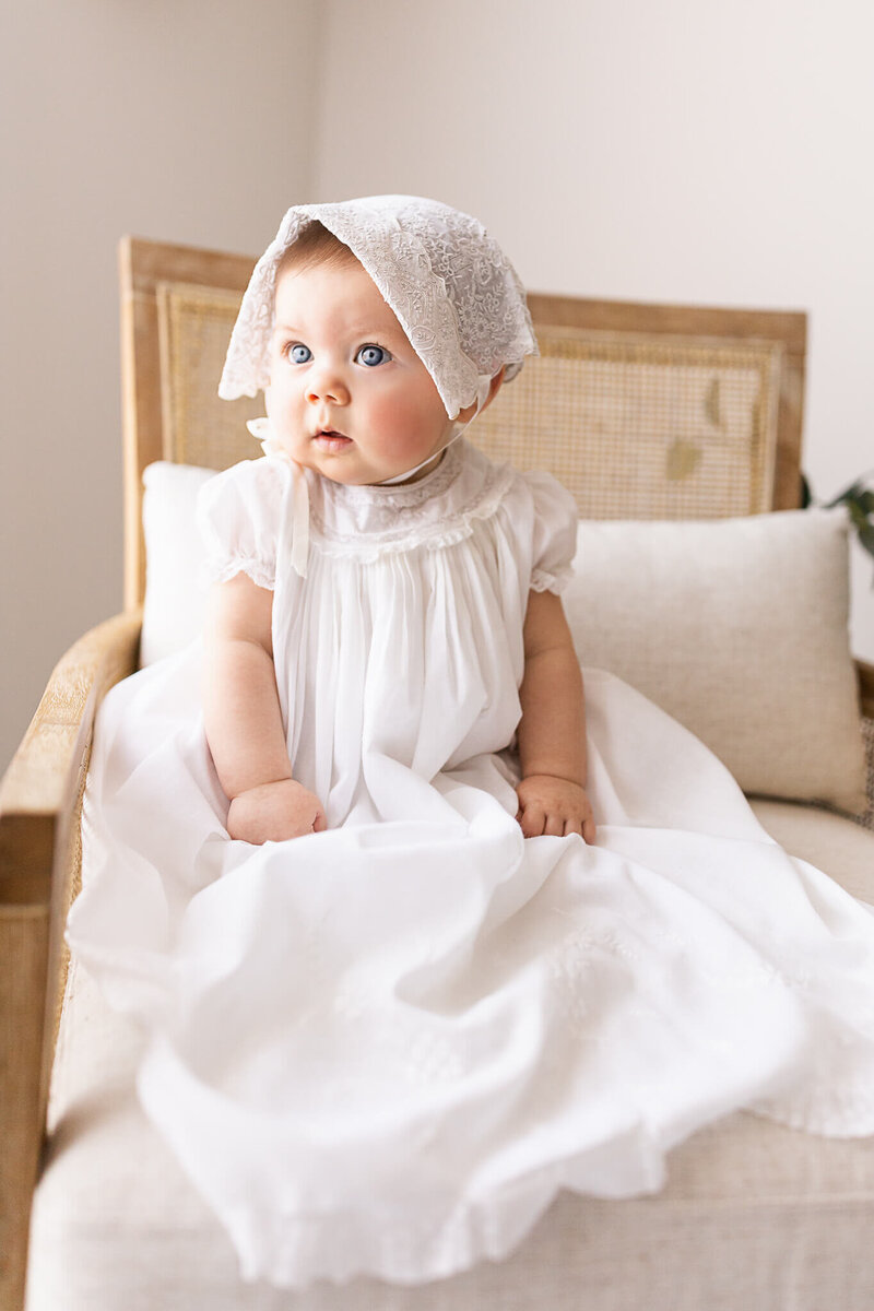 Six month baby in a white dress and white bonnet sitting in a chair in studio