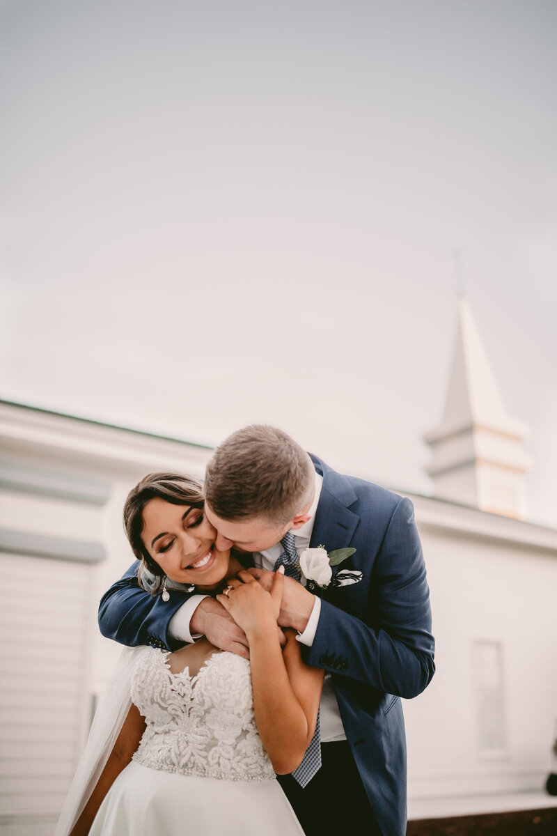 married couple standing in front of chapel, he is kissing her neck and she is smiling