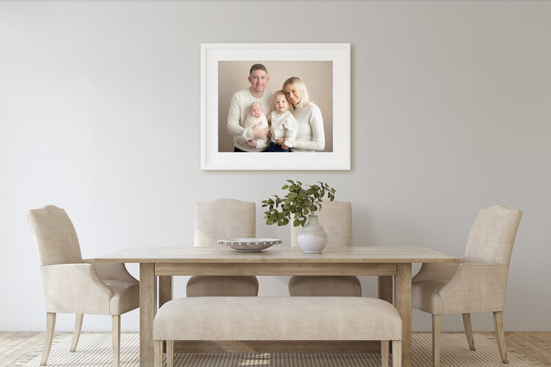 White framed photograph of a family of four with a newborn and  toddler in a dining room.
