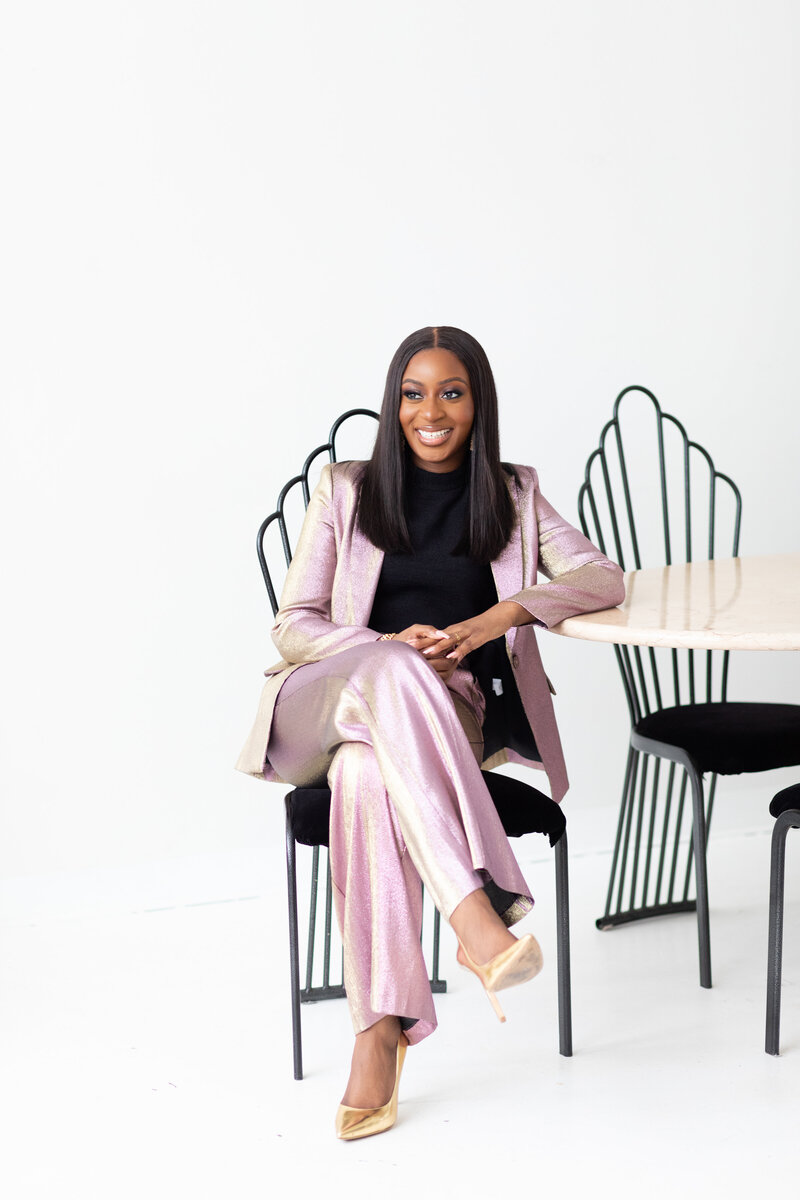 Reni, a smiling young black woman sits in on a black wire chair in a studio on a white background.