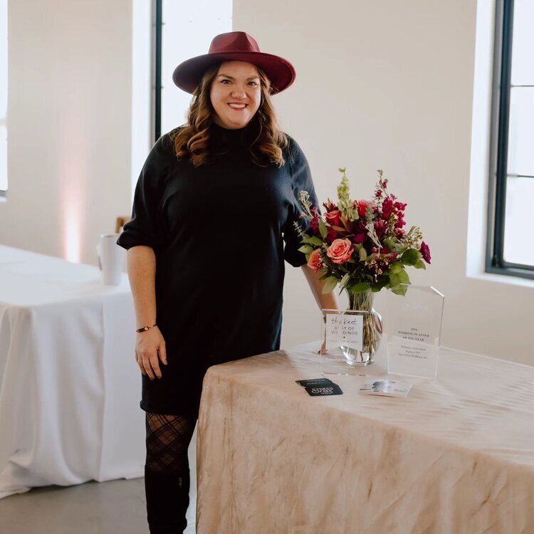 A woman in a black dress and red hat, an Iowa wedding planner, standing beside a table with floral arrangements and awards.
