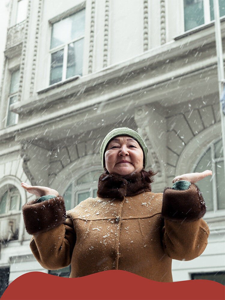 An older feminine presenting person of color smiles gently down toward the camera, lifting their hands up to touch the falling snow. They are wearing a thick, brown coat and a green winter hat. The photo is framed by a snow-like design.